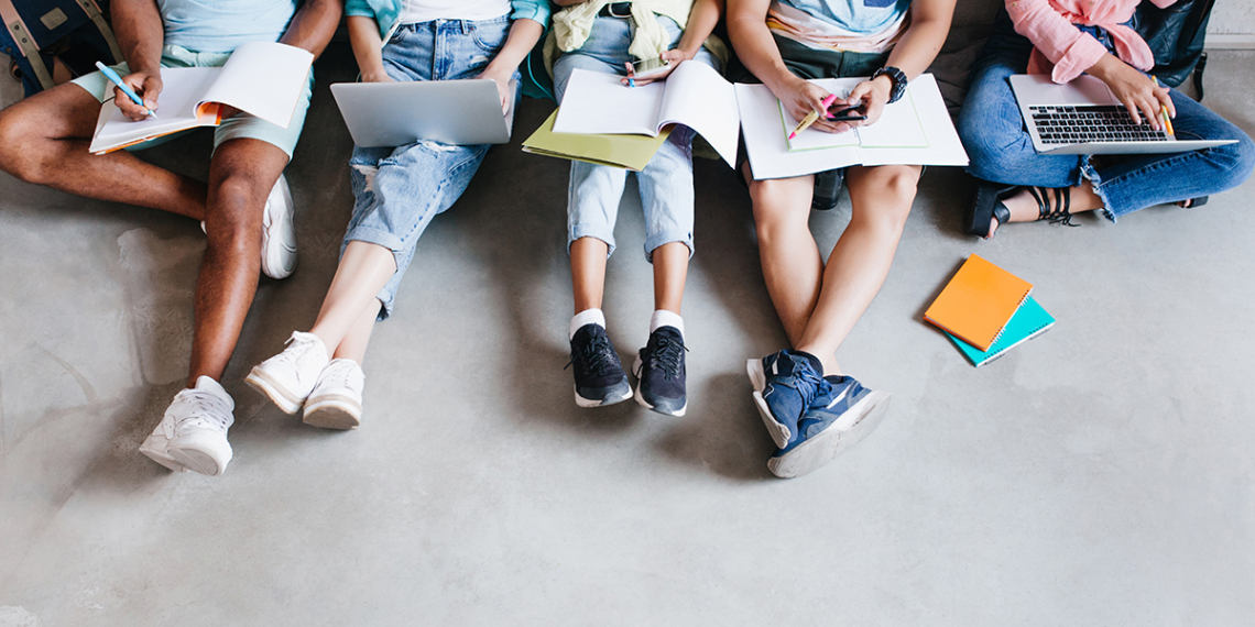 Overhead portrait of young people with laptops and smartphones, sitting together on the floor. Students writing lectures holding textbooks on their knees.
